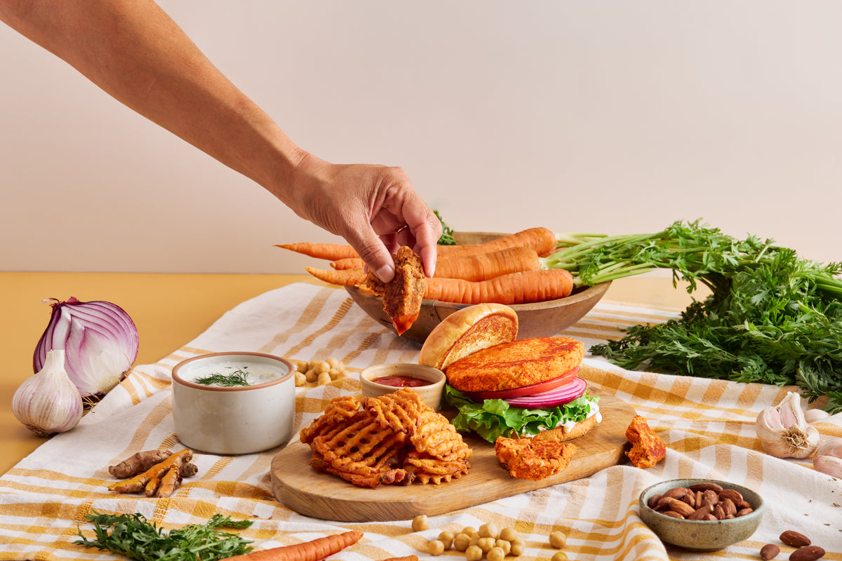 A hand dips a carrot patty beside a veggie burger, fresh carrots and almonds